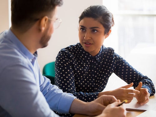 Young indian female mentor coach worker talking to male coworker teaching intern having business conversation with workmate, serious hindu manager helping colleague discussing new project in office