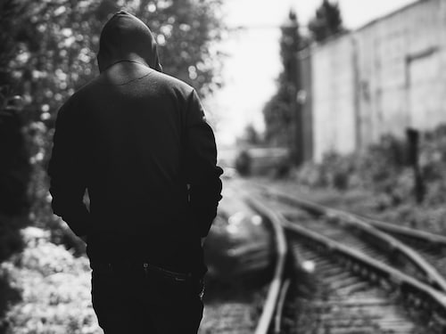 a teenager in a black hoodie walks along railway tracks in an industrial zone