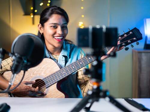 young girl influencer playing guitar during podcast or live video streaming for audience from mobile phone at home - concept of art, hobby and video blog