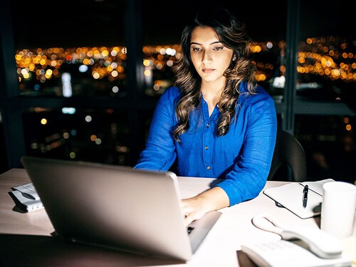 Young businesswoman working on a laptop at her office desk late into the night in front of windows overlooking the city