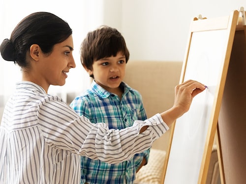 Attentive little indian boy preschooler take painting class from young millennial woman art teacher. Interested small child son watch inspired mother drawing picture on white board with colored chalks