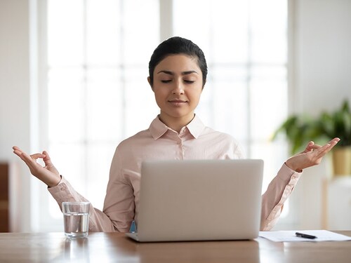 Calm young Indian businesswoman sit on desk at workplace distracted from computer job meditate in office. Peaceful mixed race female employee work on laptop practice yoga relieve negative emotions.