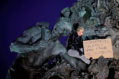 A protester holds a placard which reads "raise wages, not the retirement age" as he stands on the statue at the Place de la Nation during a demonstration against the French government"s pension reform plan in Paris as part of a day of national strike and protests in France on January 19, 2023.
