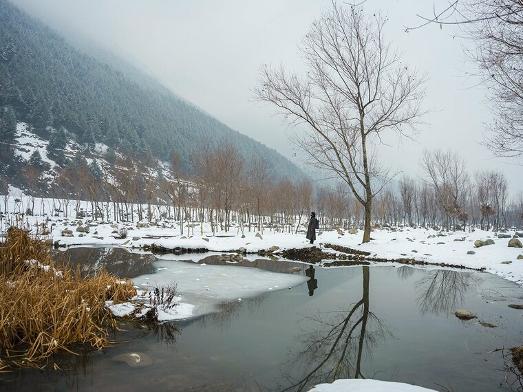 A man stands near a partially frozen pond, next to a tree, on a chilly day in Ganderbal, Jammu & Kashmir on 4th January, 2025. The India Meteorological Department (IMD) has forecasted heavy snowfall in Jammu and Kashmir on Saturday and Sunday, accompanied by light to moderate rainfall in the nearby plains. The heaviest snowfall is expected from Saturday night to Monday morning.