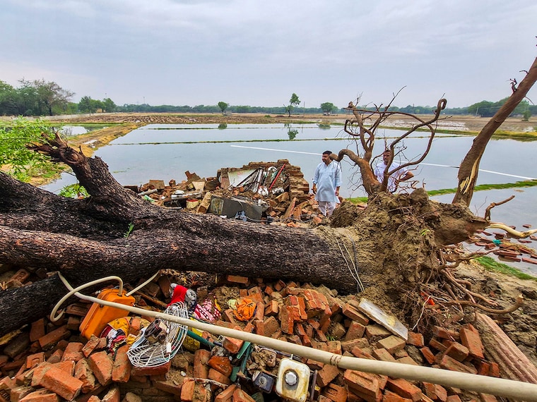 People stand near the debris of a house after a tree fell on it following strong gusty winds that accompanied rains in the city, causing it to collapse, at Najafgarh area, in New Delhi, Friday, May 2, 2025.