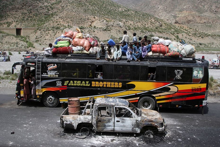 A bus with passengers sitting on the roof with belongings drives past a damaged vehicle a day after 